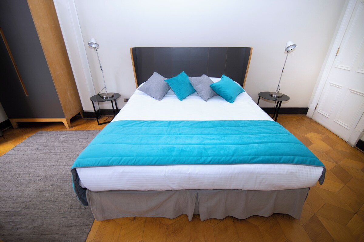 A neatly made bed with a blue blanket and grey pillows, two bedside tables with lamps, and a grey rug on a wooden floor.