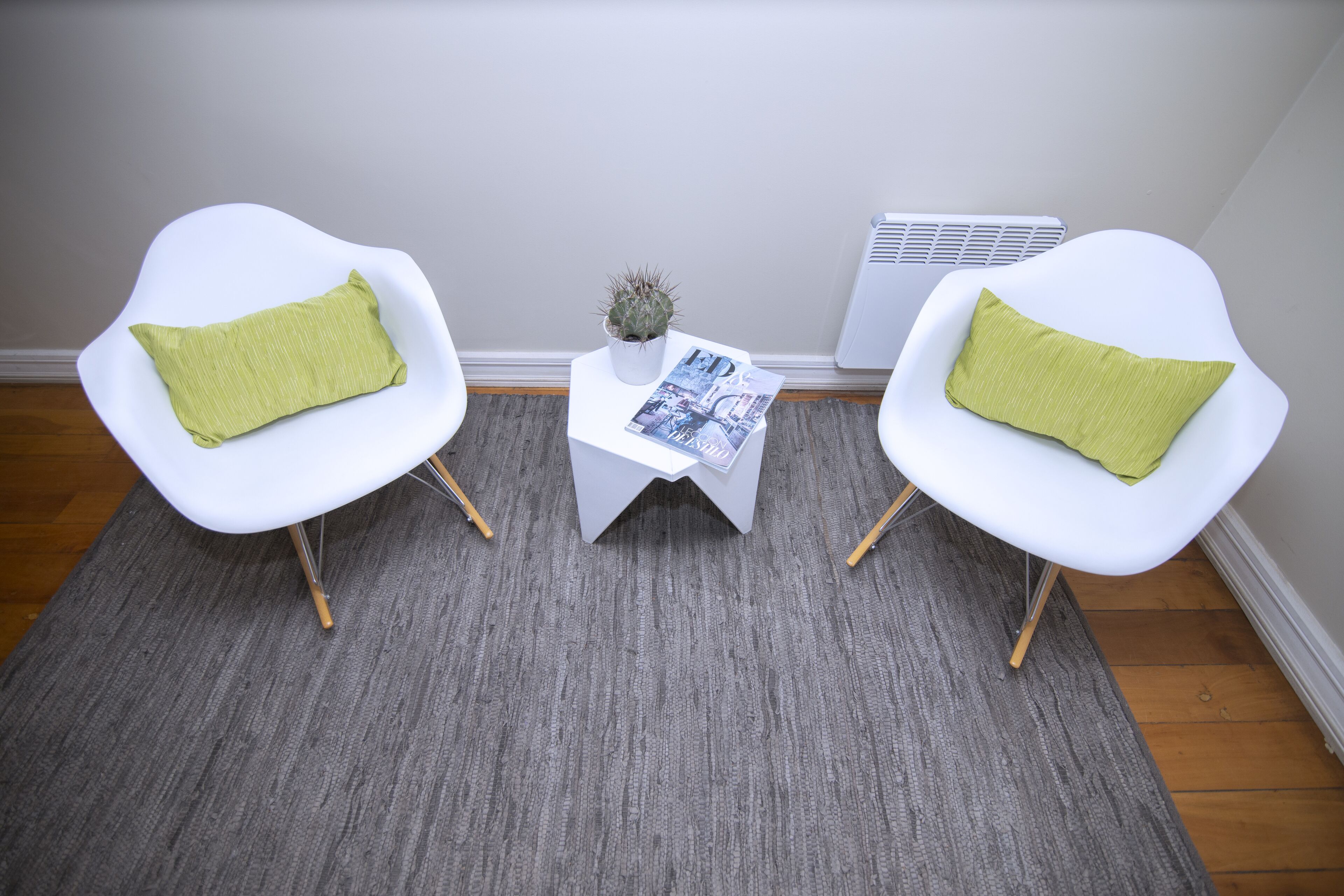 Two white chairs with green cushions, a small table with a cactus and a magazine, and an air conditioning unit against a wall.