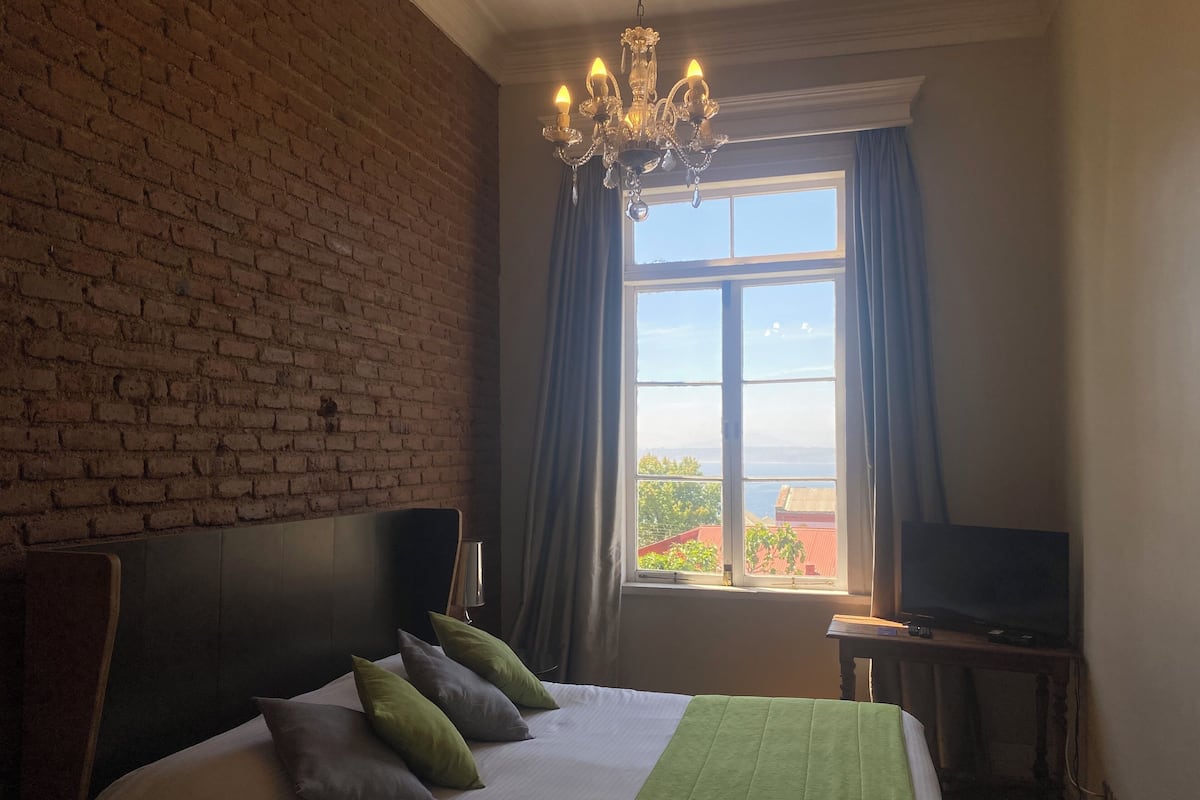 Habitación de hotel con pared de ladrillo, una cama grande con almohadas, una araña y un televisor sobre una mesita auxiliar, además de un ventanal con cortinas.