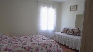 A bedroom with a floral bedspread, a small bedside table with a lamp, and a window with sheer curtains. - Casa Rural Dehesa de Algar (Algar)
