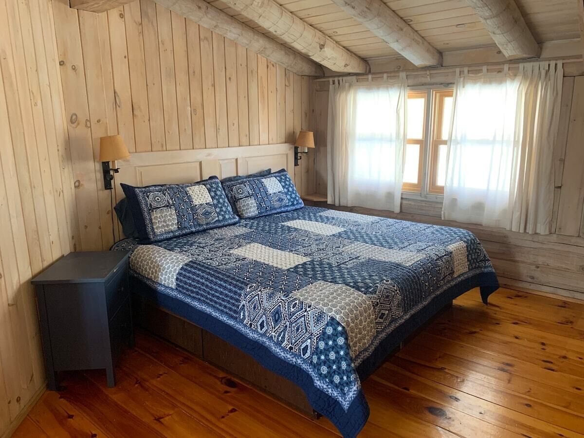 Een slaapkamer met een houten vloer, een bed met een beddeken met dessin, twee nachtlampjes en een raam met gordijnen.