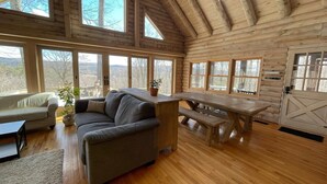 A cozy cabin interior with a wooden ceiling, large windows, a sofa, a dining table, and a plant.