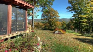 A wooden cabin with a glass enclosure, surrounded by a natural landscape with trees and a clear sky.