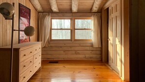 A wooden cabin room with a wooden dresser, two windows with curtains, and a wooden ceiling.