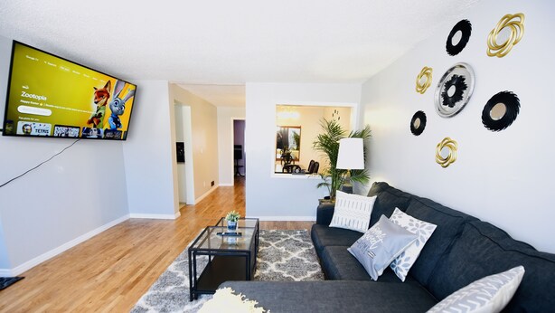 A modern living room with a flat-screen TV, a dark-colored sofa, a glass coffee table, and a patterned rug. - Prime Location Duo Nest & Rest (Kingston)