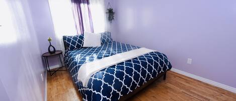 A bedroom with a bed, wooden flooring, purple walls, a window with curtains, and a small table with a plant.