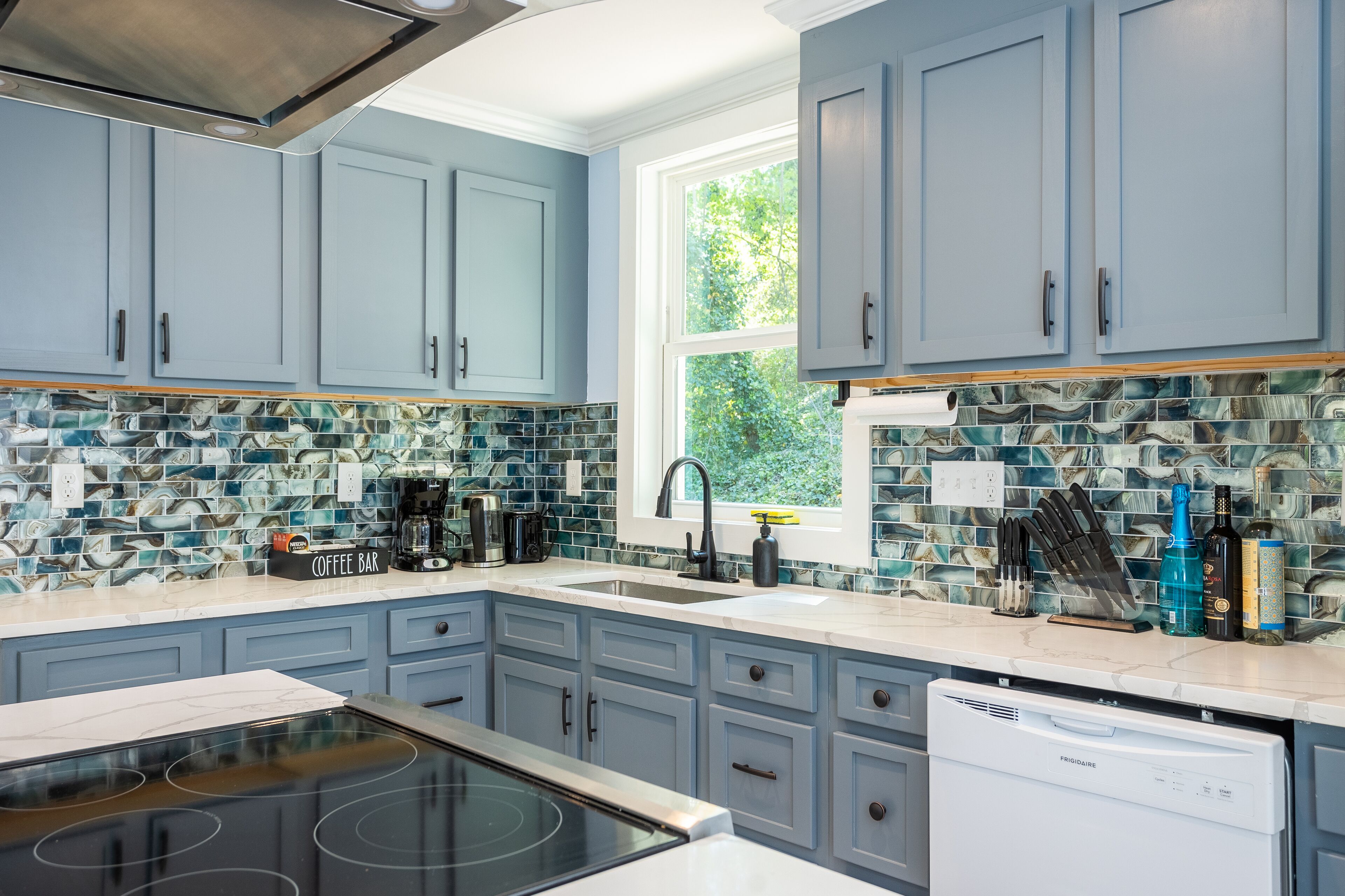 A kitchen with light blue cabinets, a large window, and a backsplash with blue and white tiles.