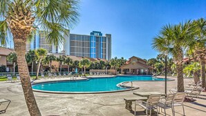 A pool area with lounge chairs, palm trees, and a hotel building in the background.