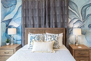 A bed with a wooden headboard, two bedside tables with lamps, patterned curtains, and a wall with abstract art. - Moana Sunrise Villa- Resort-like pool, hot tub and A/C! This place has it all! (Keaau)