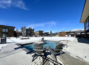 Un área de piscina con sillas y un jacuzzi, rodeado de edificios y un paisaje nevado.