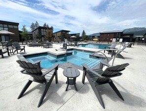 An outdoor pool area with lounge chairs and tables, surrounded by modern buildings and trees.