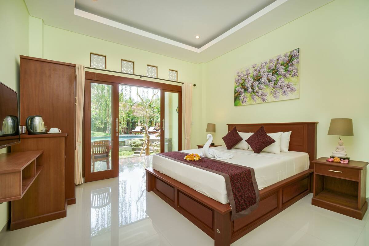 A spacious bedroom with a large bed, wooden furniture, a painting, and a view of a garden.