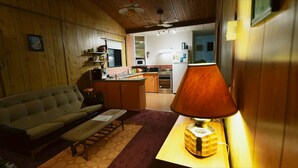 Un salon confortable avec un canapé, une table basse et une lampe. Au fond, on aperçoit une cuisine équipée d’un four à micro-ondes et d’une cuisinière.