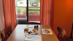 Un coin repas avec une table dressée pour deux personnes, comprenant un plateau avec de la nourriture et des condiments, et une vue sur un quartier résidentiel par la fenêtre.