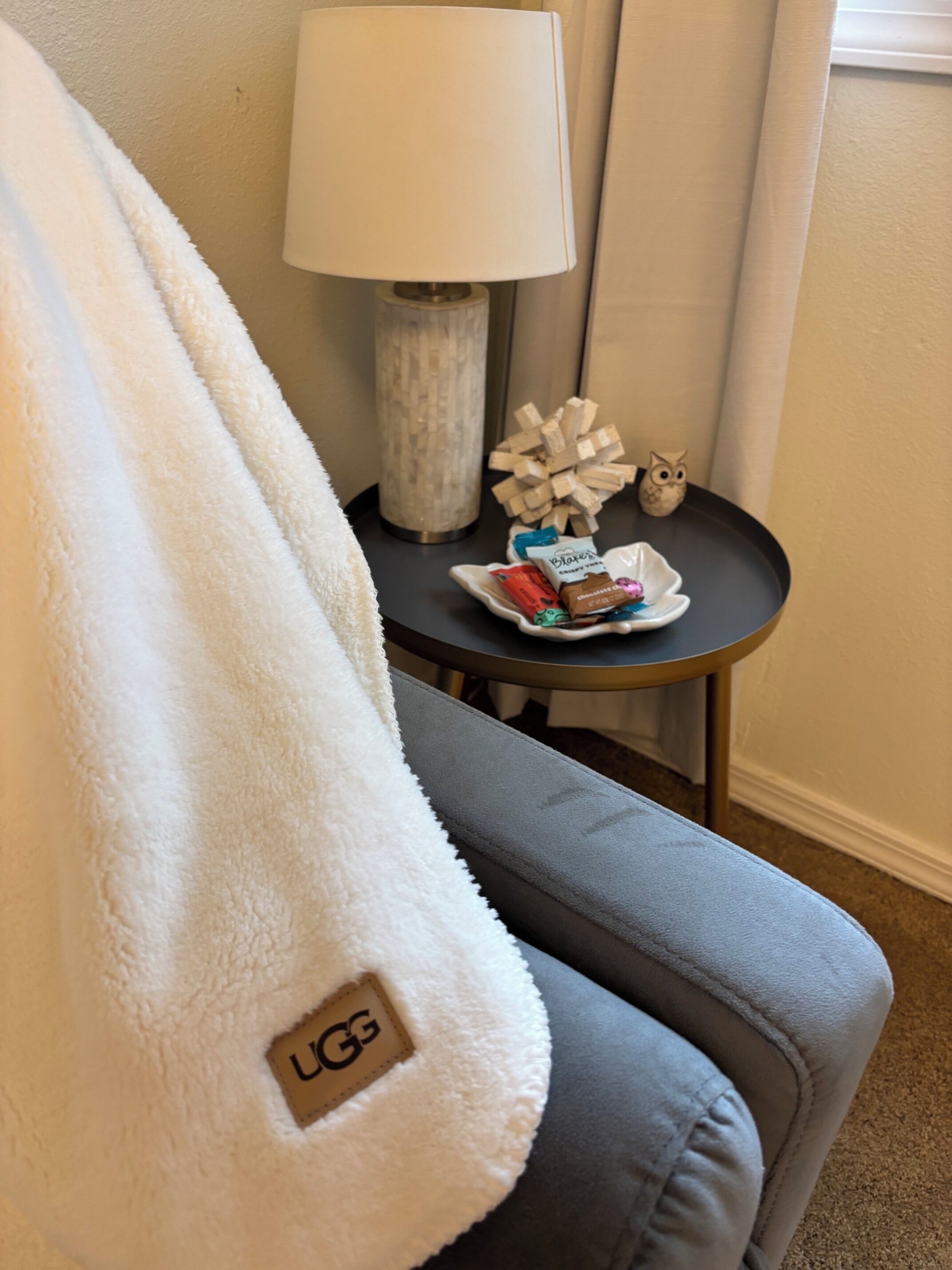 A cozy room with a grey sofa, a white fluffy blanket, a lamp, a decorative wooden sphere, and a plate with chocolates.