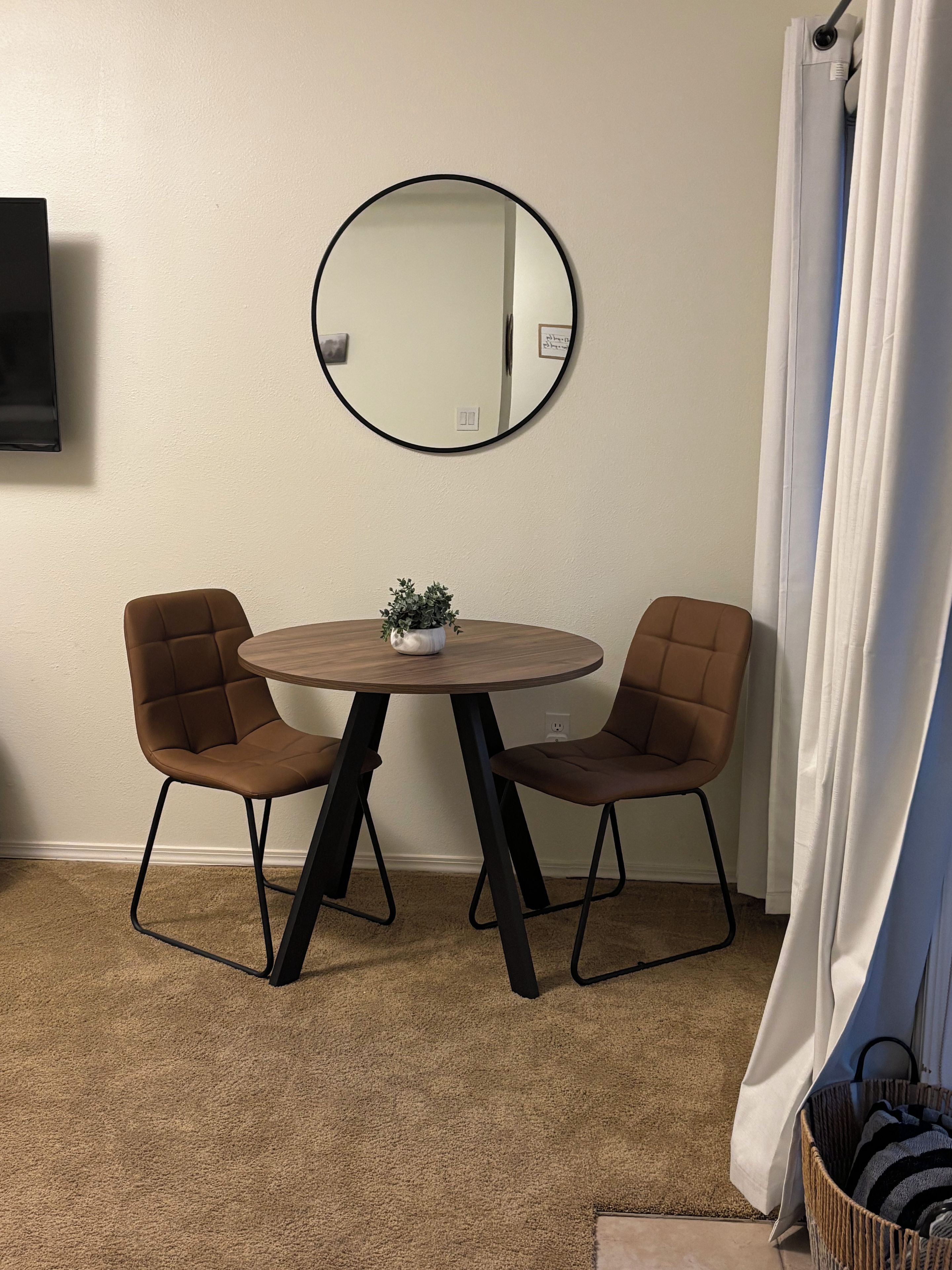 A room with a round table, two chairs, a potted plant, a wall-mounted mirror, a television, and a curtain.