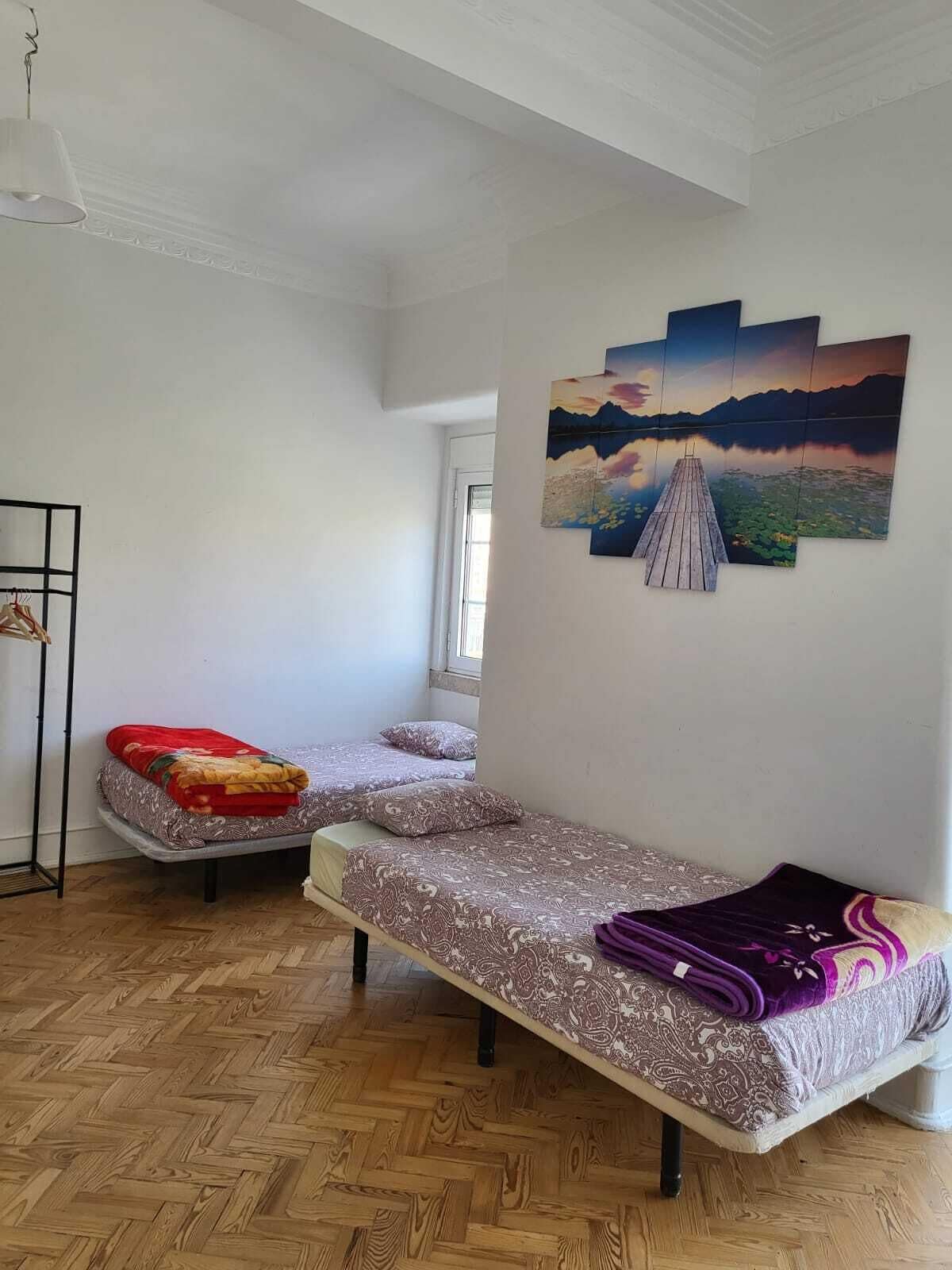 A room with two beds, a wooden floor, a painting of a lake, and a window.