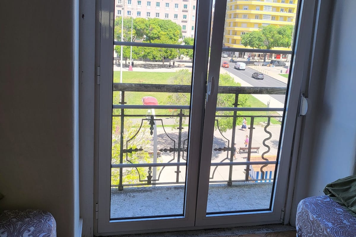 Una habitación con piso de madera en patrón de espiga, una cama con colcha estampada y una puerta corrediza de vidrio que da a un balcón exterior con vistas a edificios y a la calle.