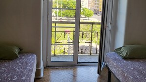 Habitación con dos camas, piso de madera en diseño espiga y puerta corredera de vidrio que da a un espacio exterior con banco y árboles.