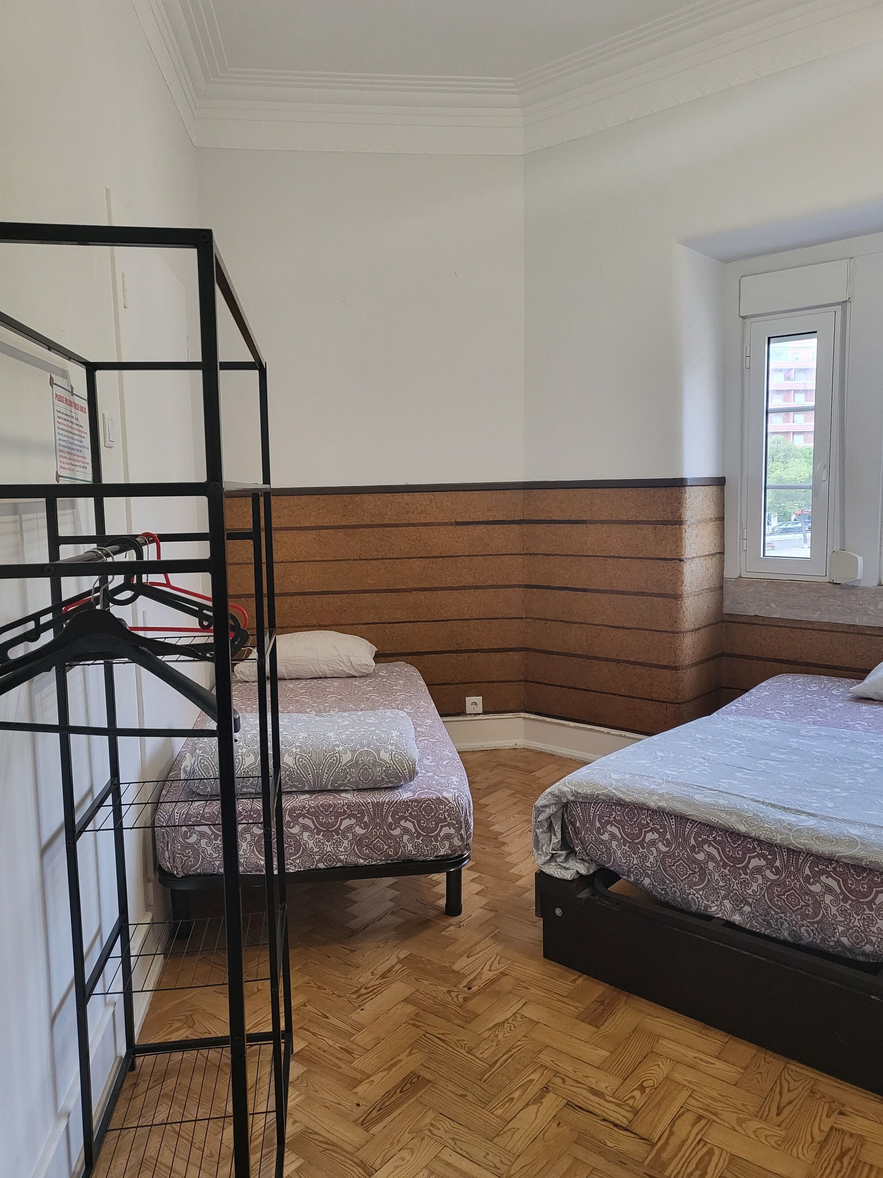 Une chambre avec deux lits, un plancher en bois, une armoire en métal noir et une fenêtre donnant sur des immeubles.