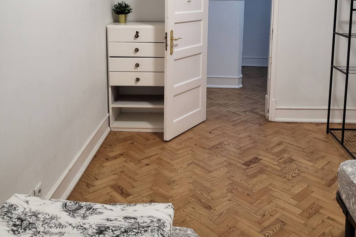 Una habitación con suelo de madera en patrón de espiga, un aparador blanco con cajones, una cama con colcha estampada y una puerta que da a otra habitación.