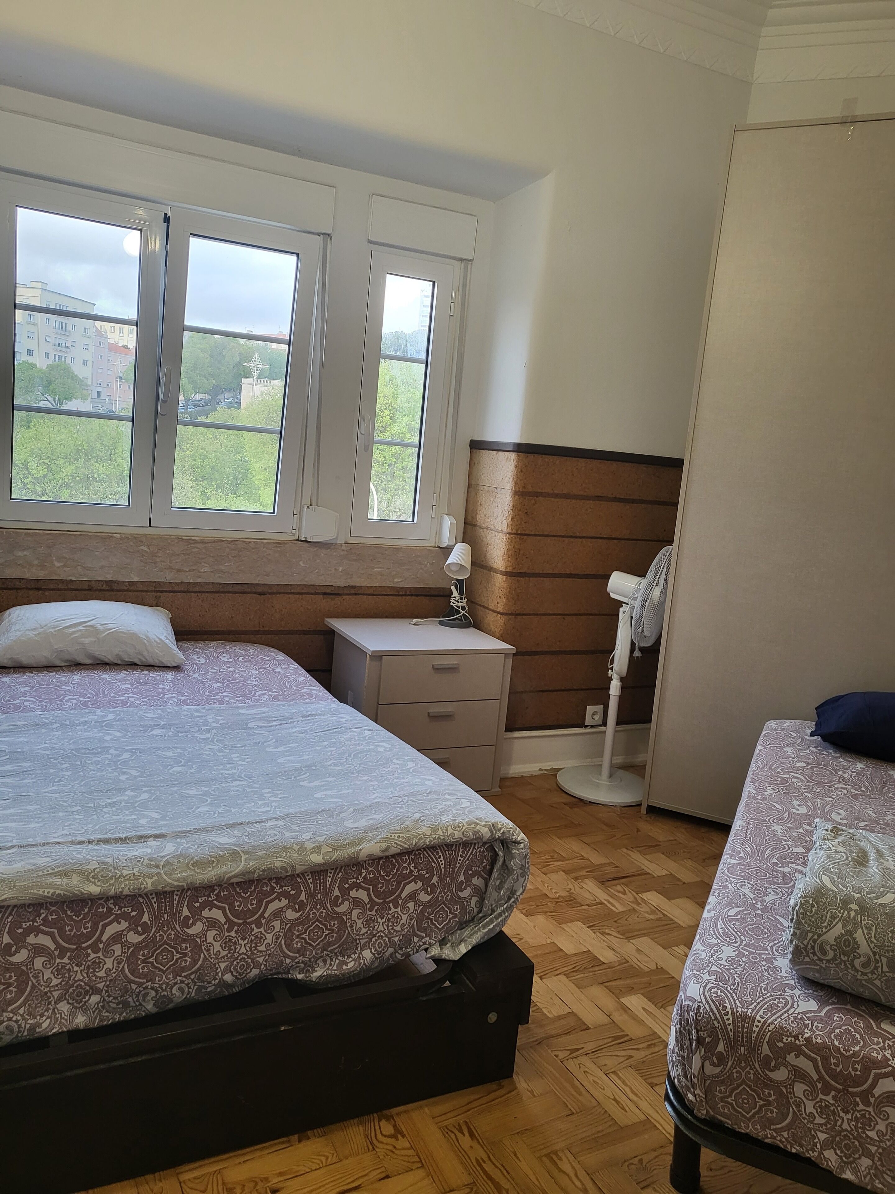 A bedroom with two beds, a nightstand, a bedside table, a standing fan, and a window with a view of buildings and trees.