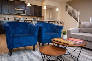 A modern kitchen with dark cabinets, a microwave, and a stove. There are two blue armchairs and a grey sofa in the living area. A small round table with a plant and a book is in the center.