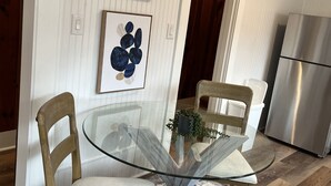 A dining area with a glass table, two wooden chairs, a white cushioned bench, and two abstract paintings on the wall.