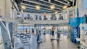 Een meerlaagse sportschool met diverse fitnessapparaten, een balkon met uitzicht, en een grote blauwe machine met de tekst 'PASANI'.