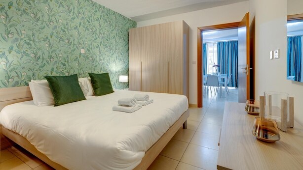 A hotel room with a large bed, a wooden wardrobe, a desk with a lamp, and a view of the dining area through an open door.
