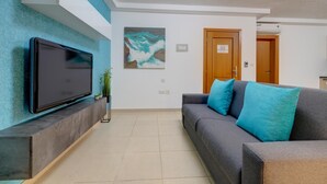 A living room with a flat-screen TV mounted on a wall, a gray sofa with blue cushions, a painting of waves, and a wooden door in the background.