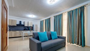 A modern living room with a grey sofa and teal cushions, a kitchen with wooden cabinets, and striped curtains.