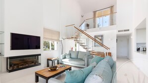 A spacious living room with a large grey sofa, a wooden coffee table, and a flat-screen TV mounted on the wall. A staircase with a glass railing leads to an upper level.