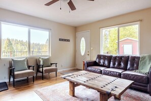 A living room with a brown leather couch, two chairs, a wooden coffee table, and a large window with a view of trees and a red building. - A rejuvenating cabin in the woods (Pioneer)