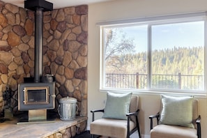 A stone fireplace with a chimney, a window with a view of a forest, and two chairs with cushions.