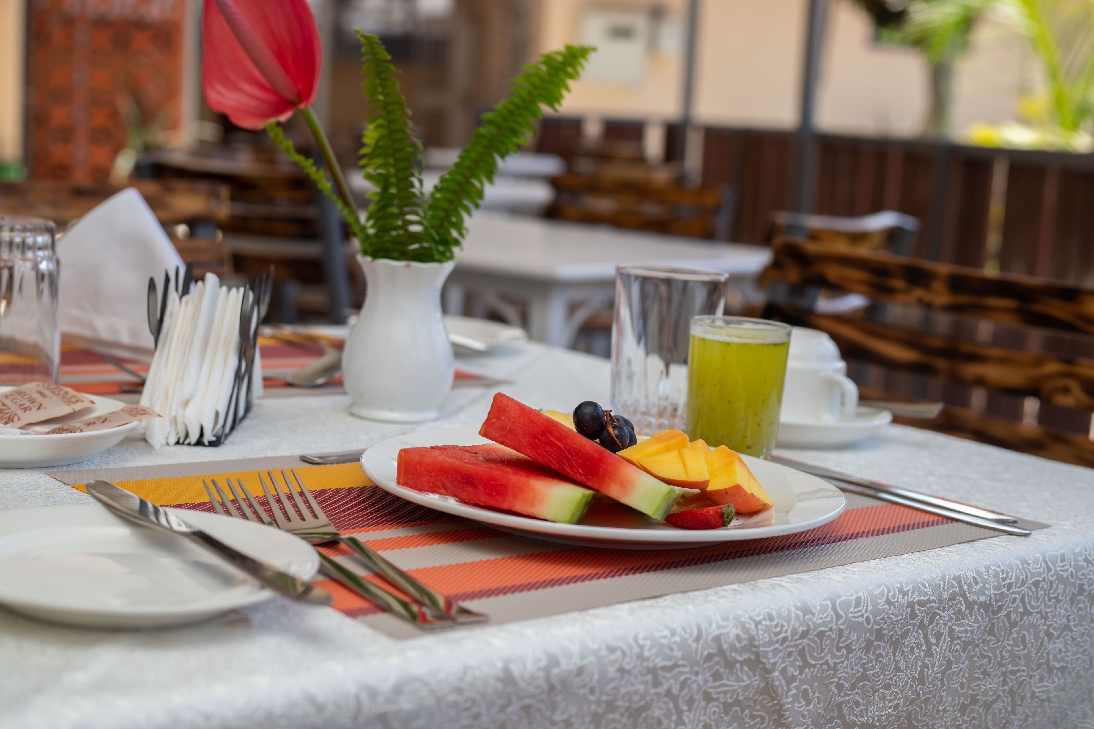 Une table mise pour le petit-déjeuner avec une assiette de fruits, un verre de jus et un vase avec une plante.