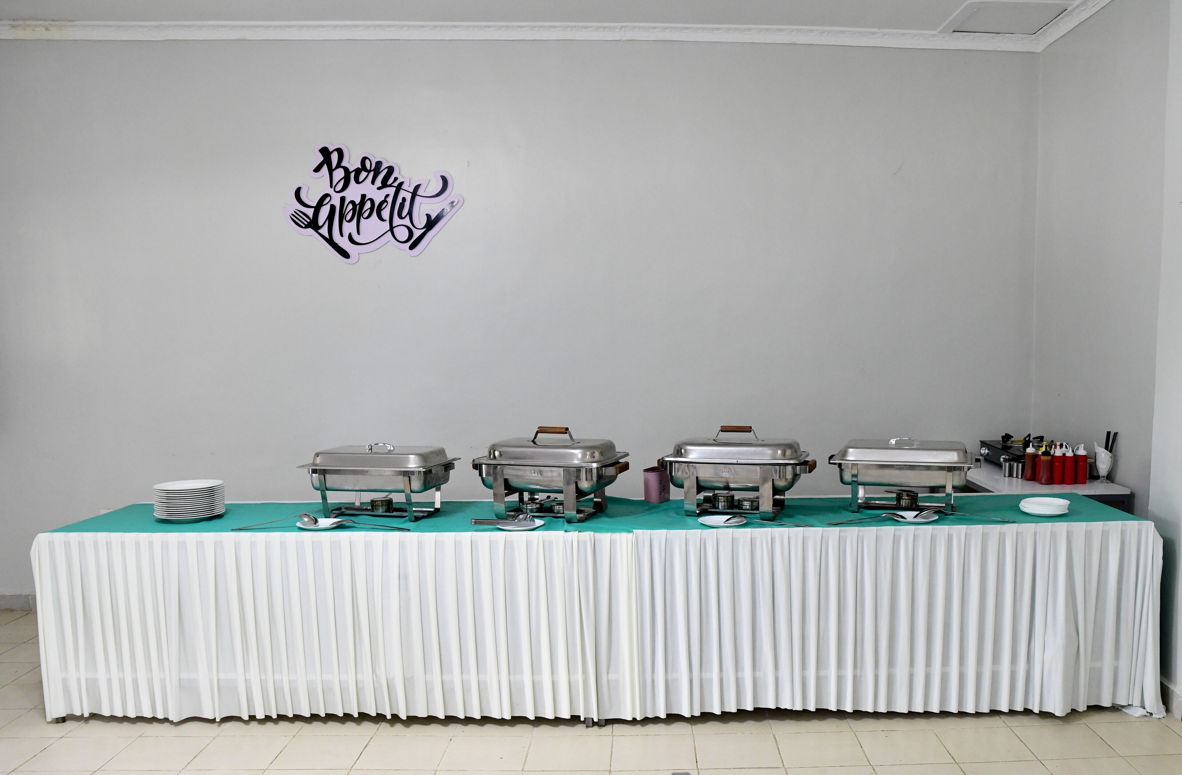 A buffet table with multiple food containers, plates, and utensils, set against a white wall with a decorative sign.