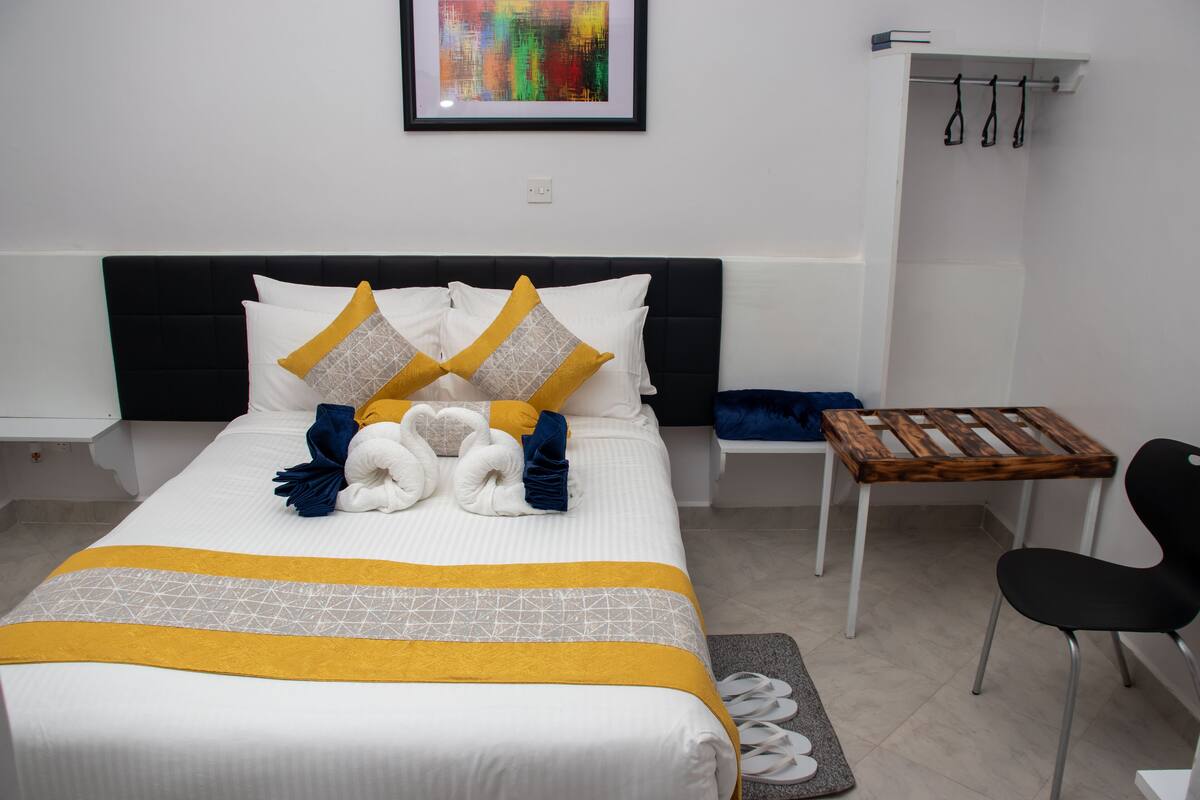 A neatly made bed with a yellow and white bedspread, a black headboard, and a small wooden table with a chair beside it.