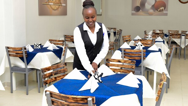 Une salle à manger avec des tables recouvertes de nappes bleues et blanches, des chaises en bois, et une personne qui dispose la vaisselle.