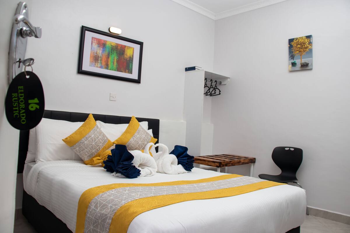 A hotel room with a neatly made bed, a small wooden side table, a black chair, and a wall-mounted coat rack.