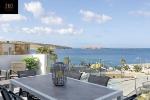 Terraço com mesa e cadeiras, oferecendo vista para a costa, céu claro e uma ilha ao longe.