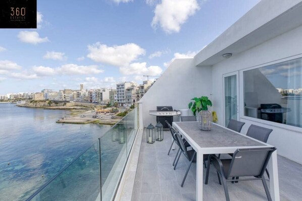 Uma varanda moderna com mesa de jantar, cadeiras e churrasqueira. Oferece vista para um corpo d'água e para a paisagem urbana.