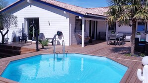 Ett område vid poolen med ett trädäck, en vit byggnad med ett tak täckt med tegelpannor, utemöbler och en palm.