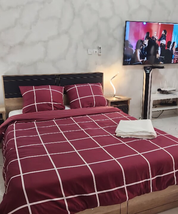 Una cama bien hecha con una colcha de cuadros rojos y blancos, un cabecero de madera y un televisor en la pared que muestra a un grupo de personas.