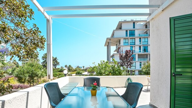 A glass-top table with four chairs, a potted plant, and a green door on a balcony with a clear view of a modern building in the background.