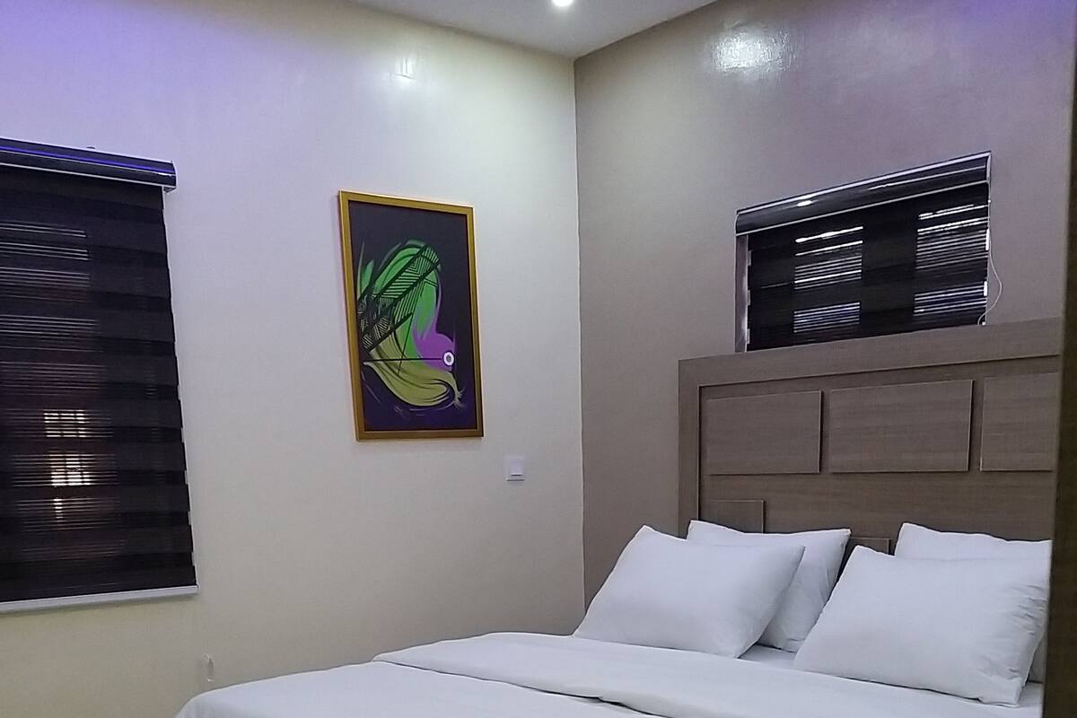 A bedroom with a bed, a headboard, a window with blinds, and a wall-mounted artwork.