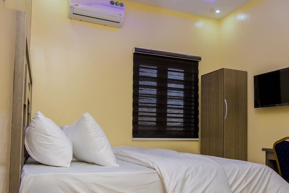 A hotel room with a bed, a desk with a telephone, a wardrobe, a television, and an air conditioning unit.