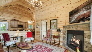 A cozy cabin interior with a fireplace, wooden walls, a dining area, and a kitchen.