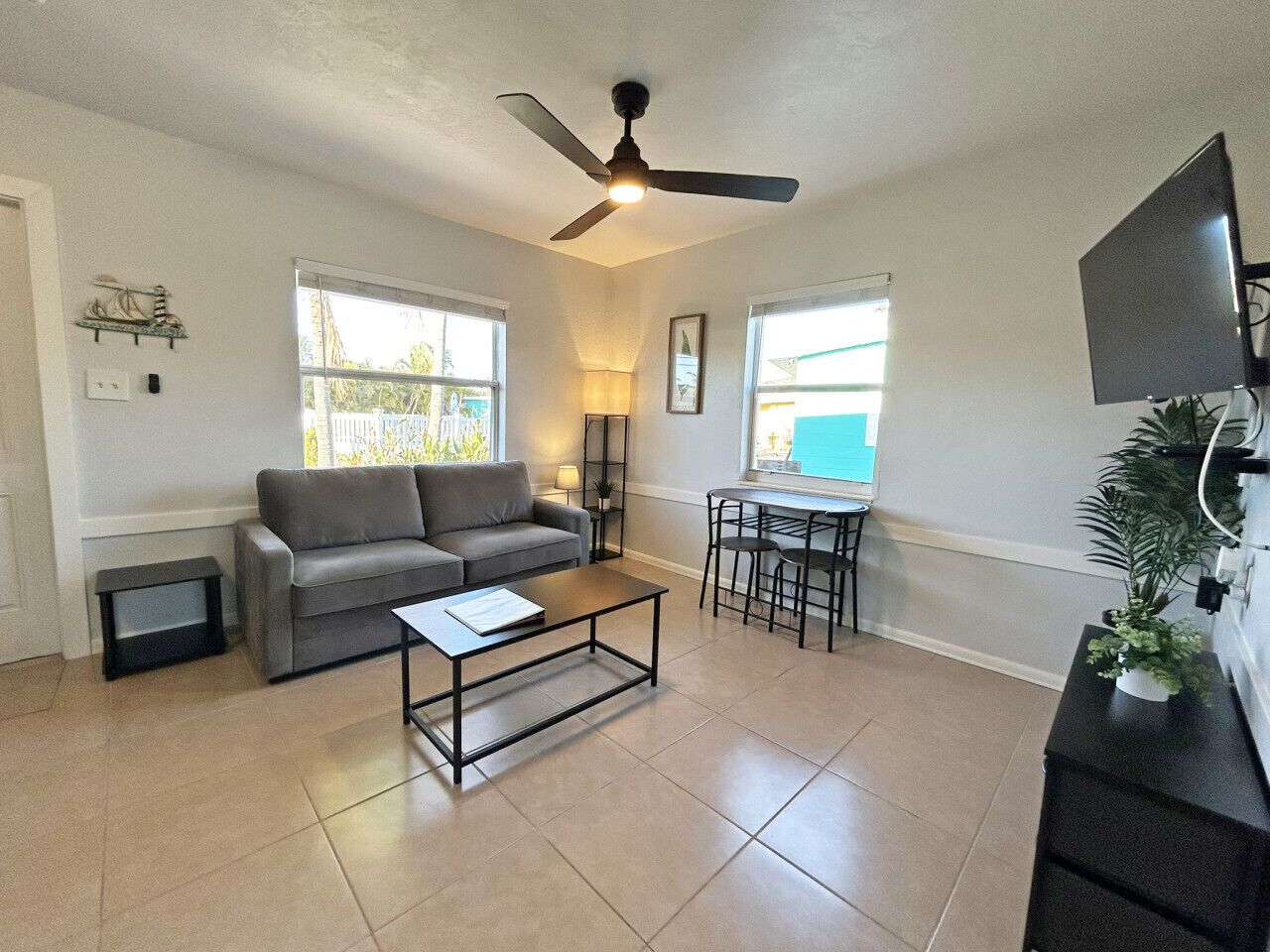 A living room with a gray sofa, a coffee table, a dining table with chairs, a ceiling fan, and a TV mounted on the wall.