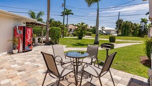 Eine Terrasse mit Tisch und Stühlen, einem Coca-Cola-Kühler und einem gepflegten Rasen mit Palmen.